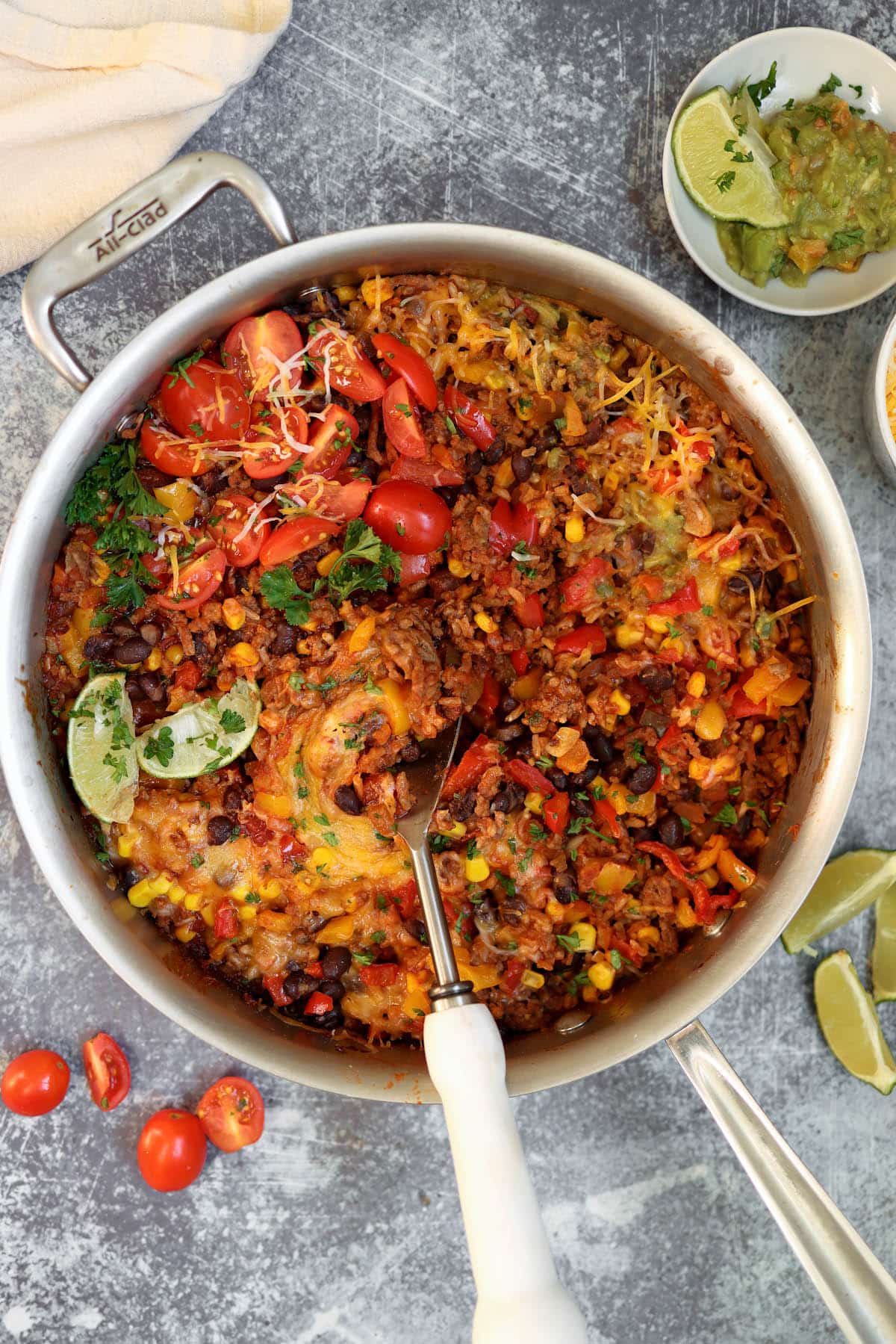 Mexican Beef and Rice skillet with tomatoes and cheese, corn and salsa on a counter top with limes, guacamole and tomatoes