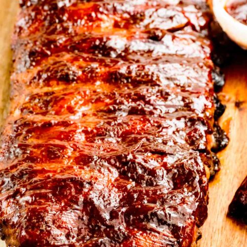 A close-up of a rack of glazed barbecue ribs on a wooden cutting board, with a small bowl of barbecue sauce partly visible in the corner.
