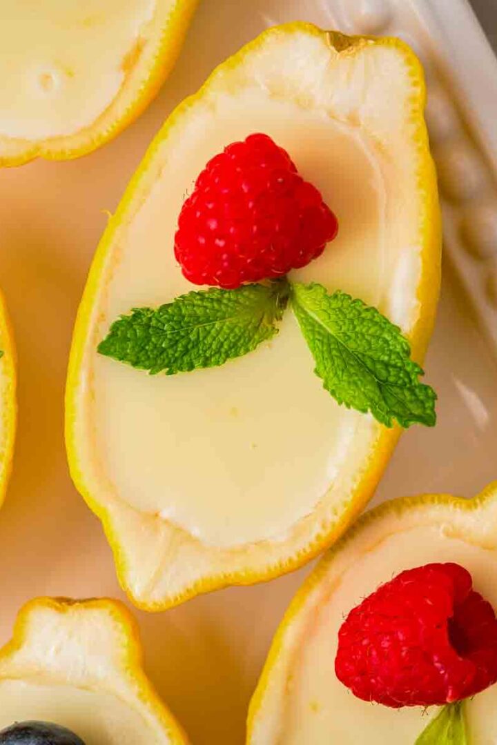 Lemon possets served in hollowed lemon cups, topped with fresh raspberries, blueberries, and mint on a white platter.