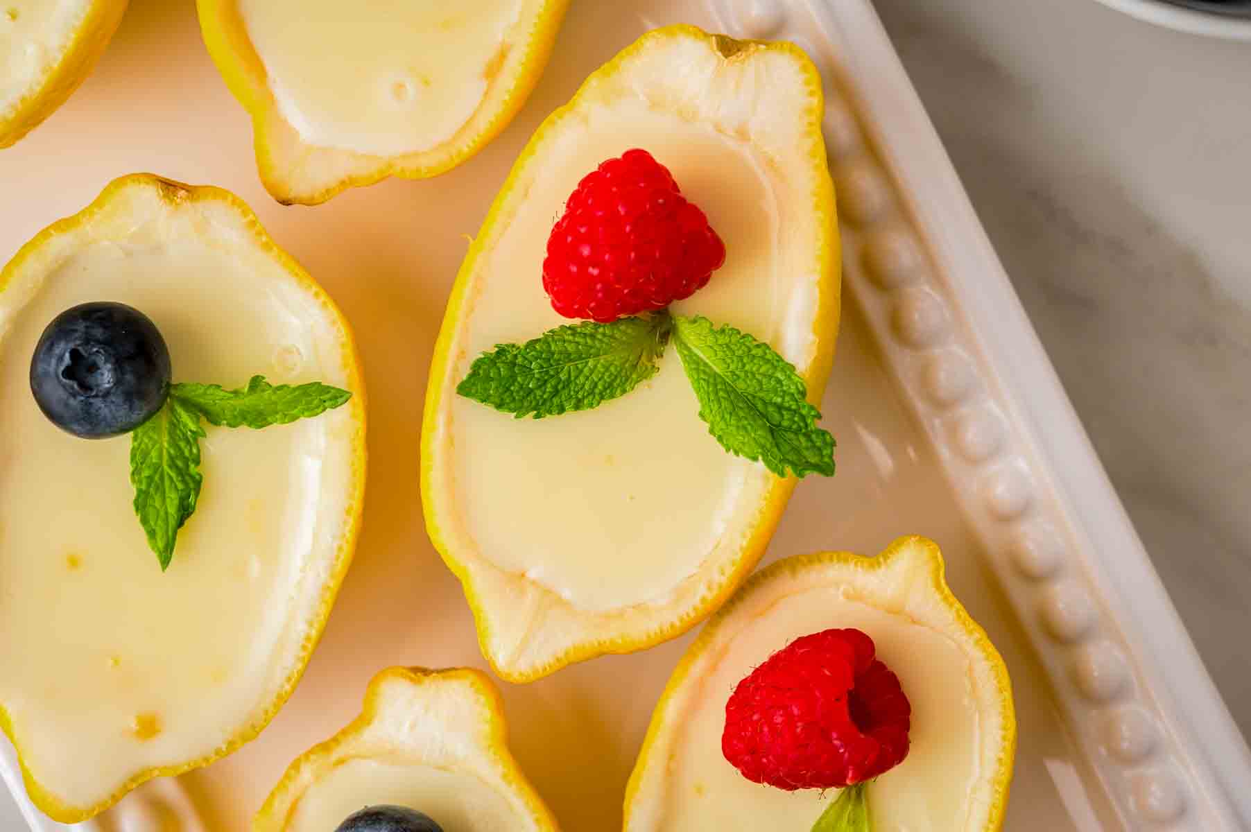 Lemon possets served in hollowed lemon cups, topped with fresh raspberries, blueberries, and mint on a white platter.