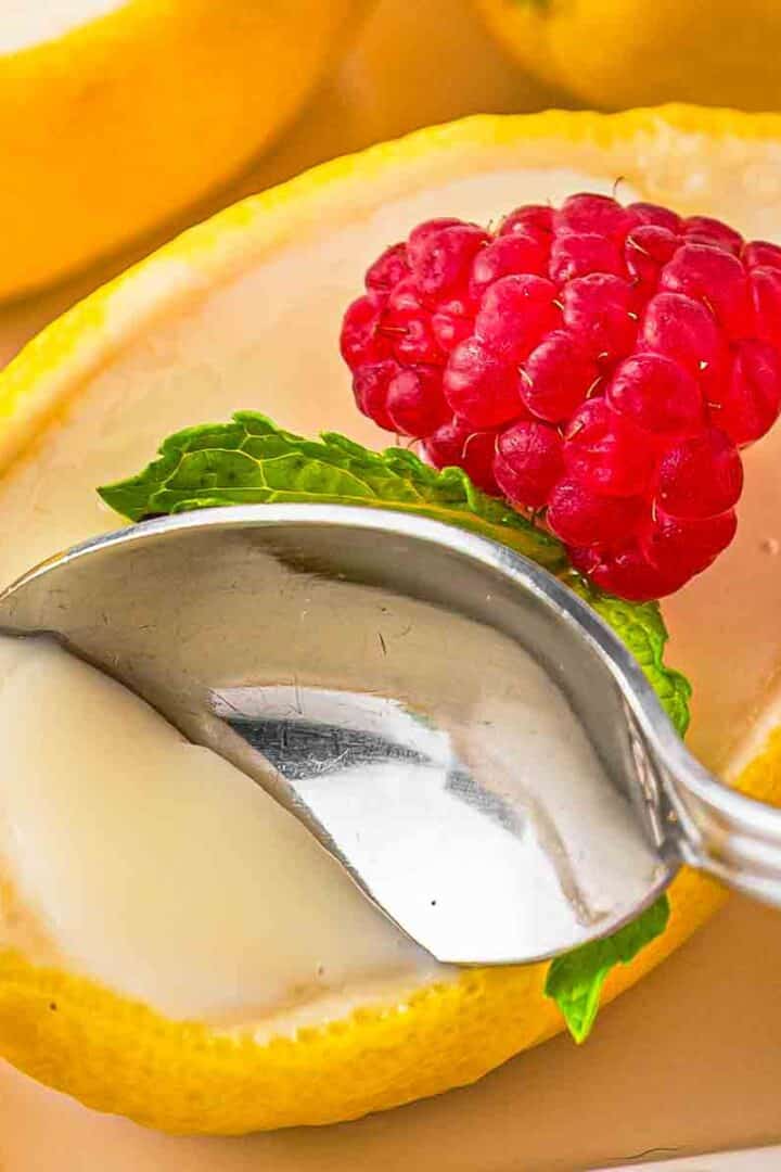 Close-up of a spoonful of creamy lemon posset with raspberry on top lifted from a lemon shell, showing smooth texture and tiny specks of zest.