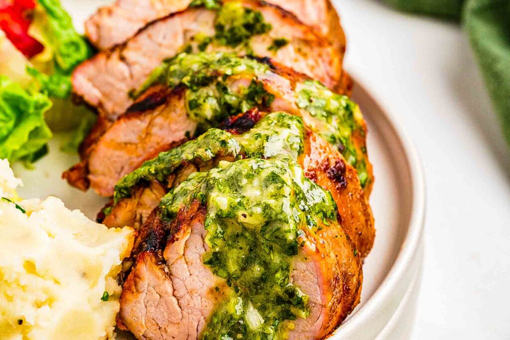 Sliced grilled pork tenderloin arranged on a platter, topped with spoonfuls of green herb sauce.