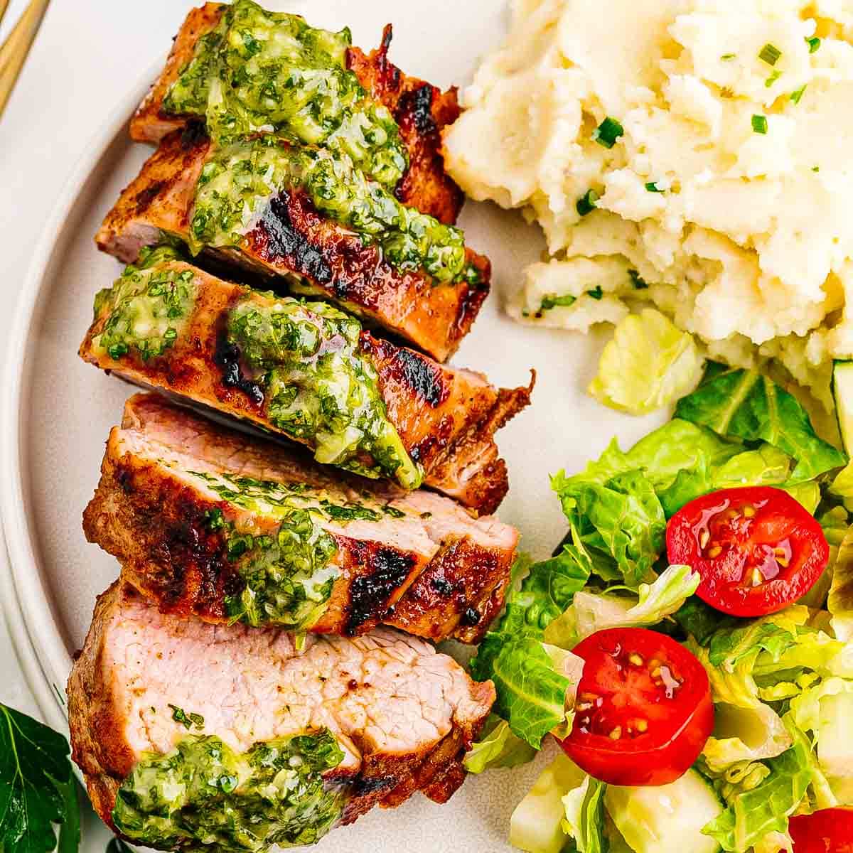 Overhead shot of a summer dinner table with grilled pork tenderloin, herb sauce, and several side dishes.