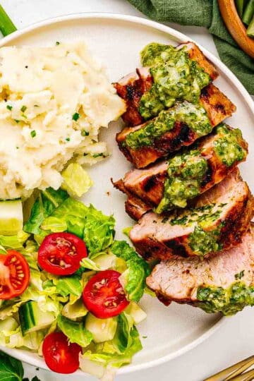 Plate of grilled pork tenderloin with herb sauce, served with a side of potatoes and a simple salad.