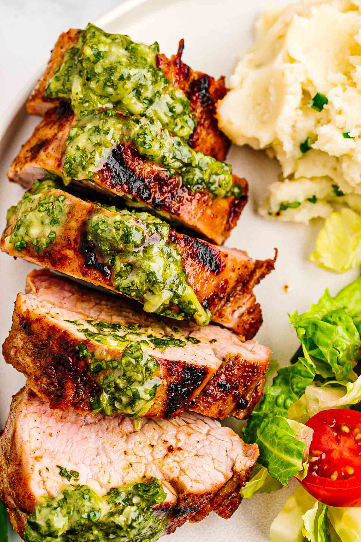 Whole grilled pork tenderloins on a cutting board with grill marks, a small bowl of herb sauce, and a knife nearby.