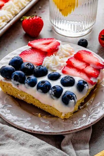 lose-up of a slice of fruit pizza showing the soft sugar cookie crust, thick Greek yogurt cream cheese frosting, and glossy berries arranged in red, white, and blue stripes