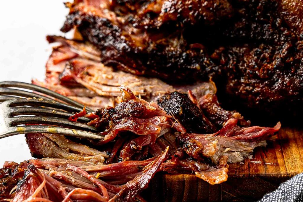 Smoked Boston butt pulled apart into tender shreds on a wooden cutting board with bark pieces and cooking juices