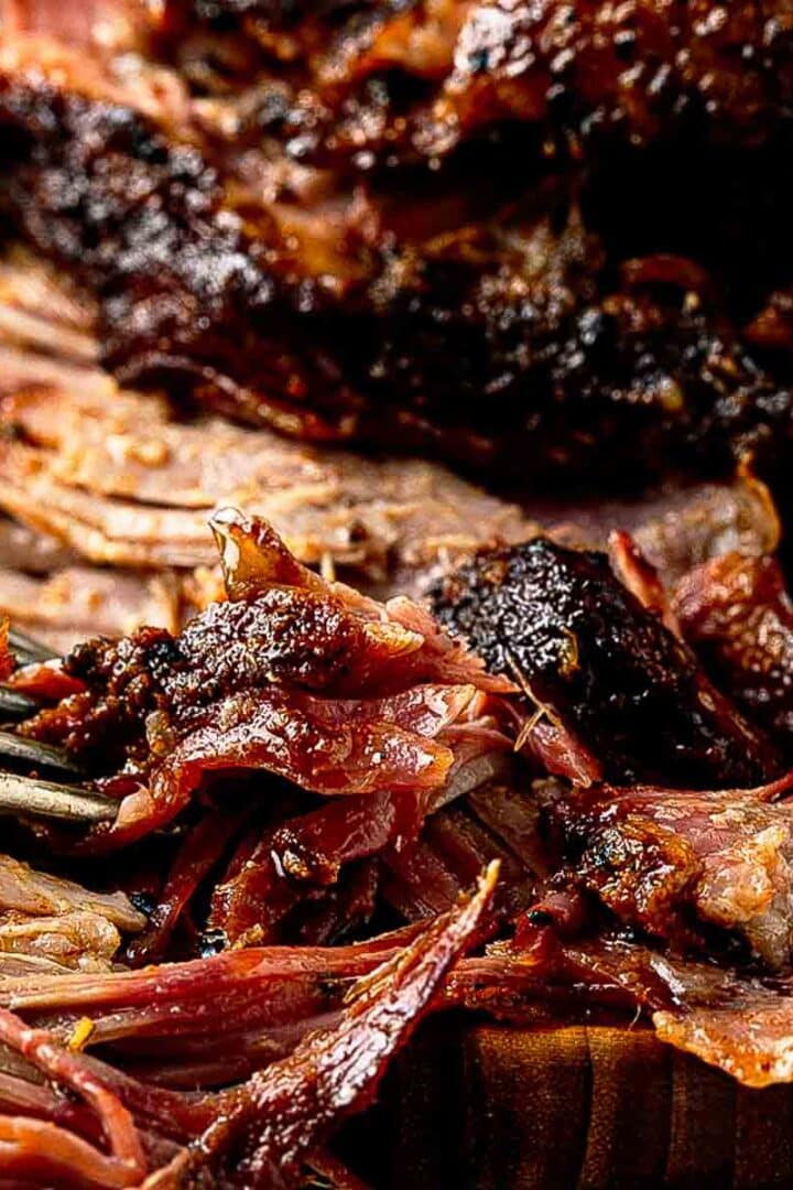 Smoked Boston butt pulled apart into tender shreds on a wooden cutting board with bark pieces and cooking juices