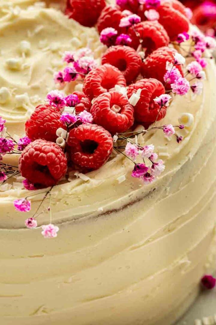 Raspberry white chocolate layer cake decorated with fresh raspberries, white chocolate curls, and spring flowers on a white cake stand