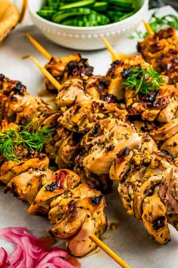 Single chicken souvlaki skewer held above a platter, showing charred edges and juicy pieces, with blurred pita and salad in the background.”