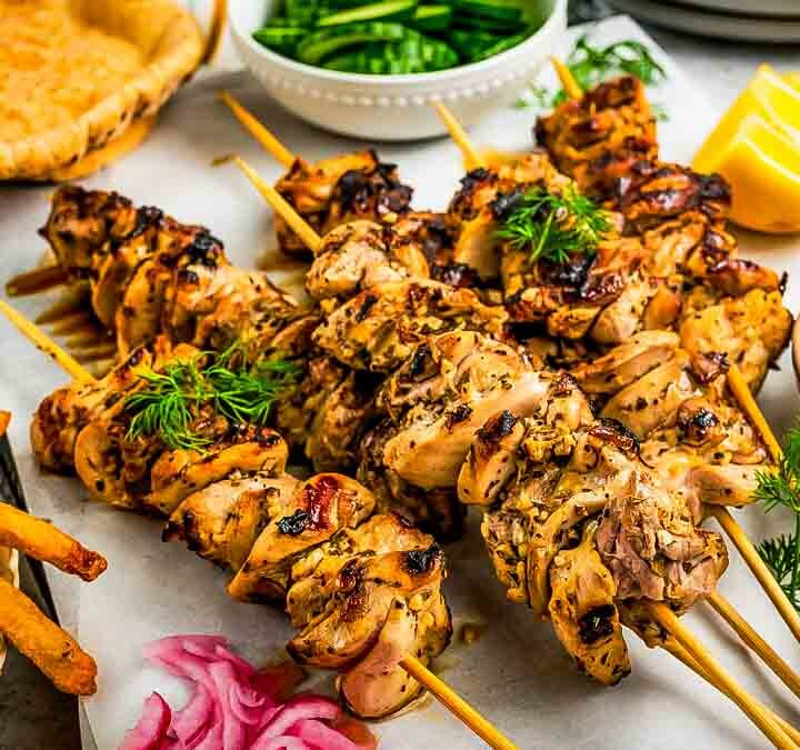 Single chicken souvlaki skewer held above a platter, showing charred edges and juicy pieces, with blurred pita and salad in the background.