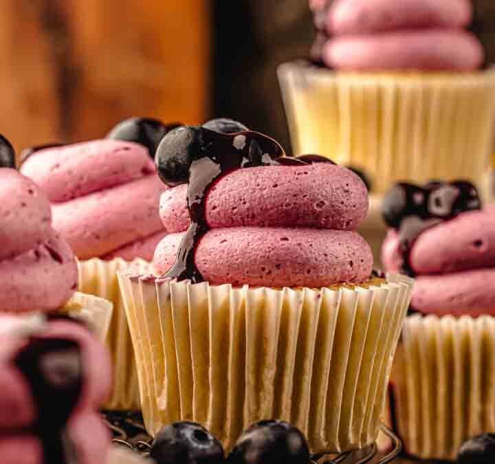Lemon blueberry cupcakes styled on a wooden board with lemons and blueberries scattered nearby.