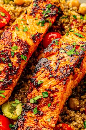 Family-style skillet of harissa salmon and quinoa on a wooden table with serving spoons nearby.