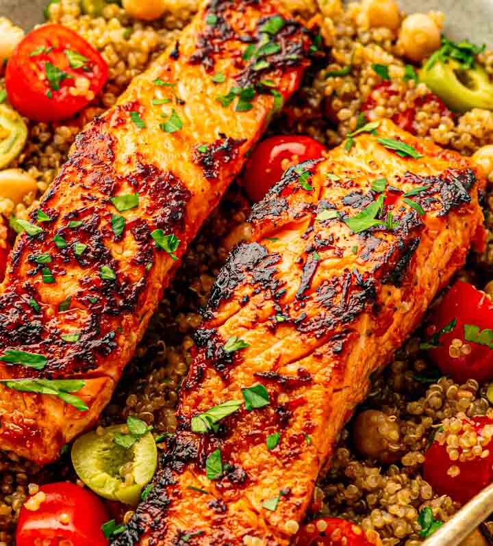 Family-style skillet of harissa salmon and quinoa on a wooden table with serving spoons nearby.
