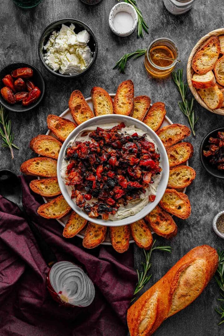 Overhead view of whipped goat cheese dip with bacon and dates in a serving dish with toasted bread on the side
