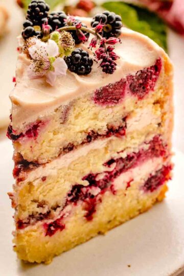 A close-up of a big slice of Blackberry layer cake with jam-streaked buttercream, fresh blackberries and edible flowers on top