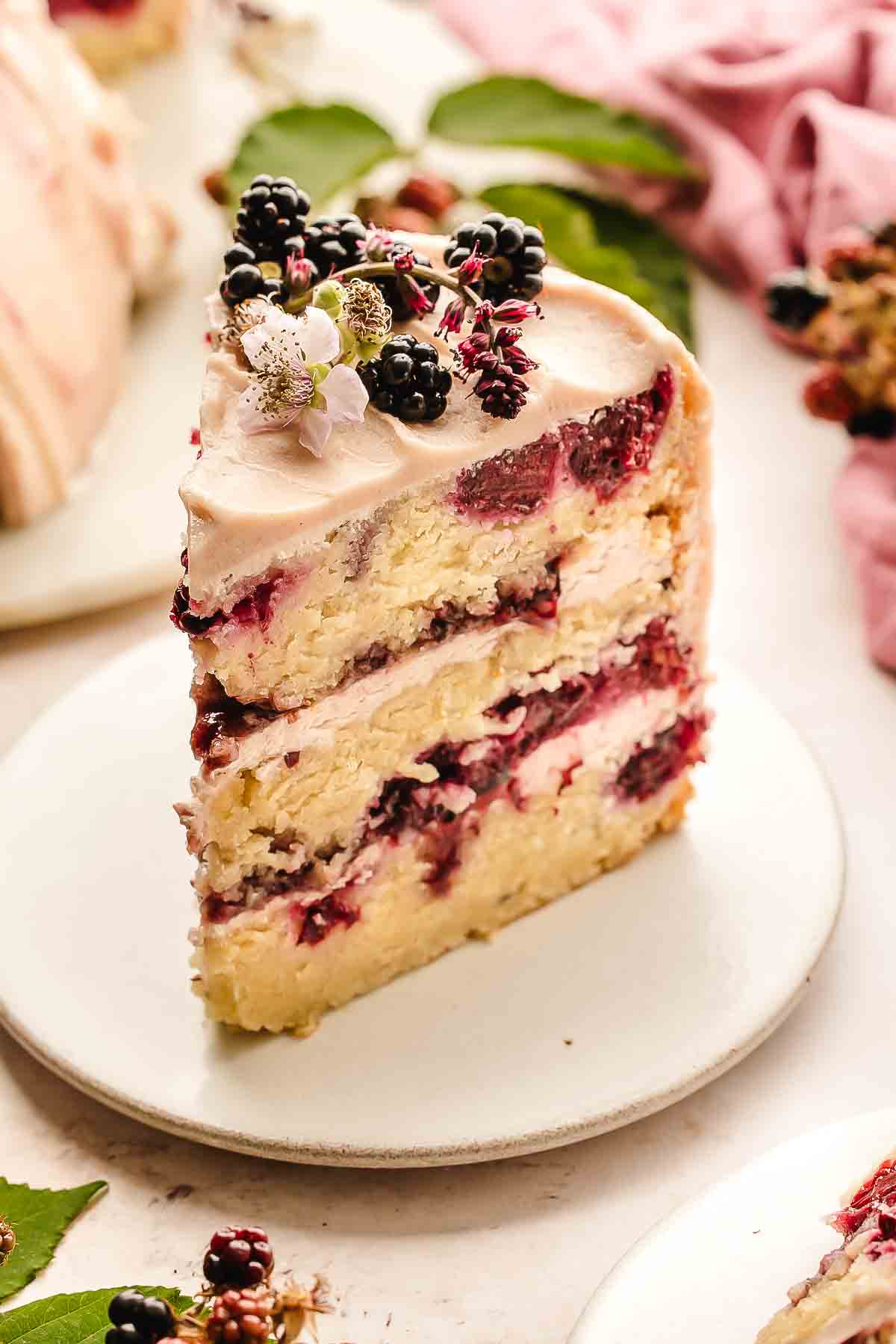 A close-up of a big slice of Blackberry layer cake with jam-streaked buttercream, fresh blackberries and edible flowers on top