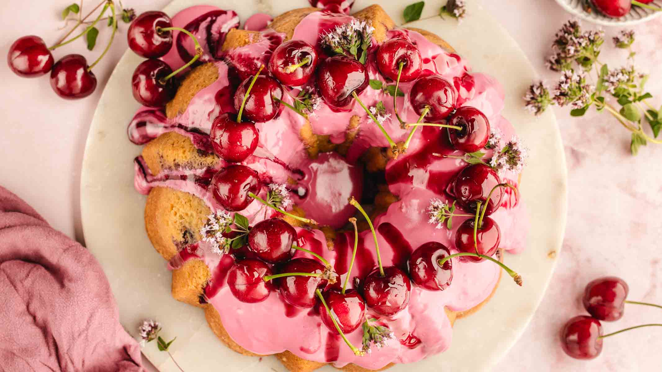 "Fresh cherry bundt cake on a cake stand, fully iced and decorated with cherries and flowers"