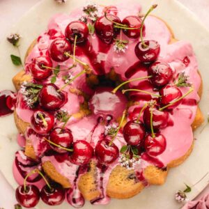 Fresh cherry bundt cake with pink cherry icing dripping down the ridges, topped with whole fresh cherries and flowers