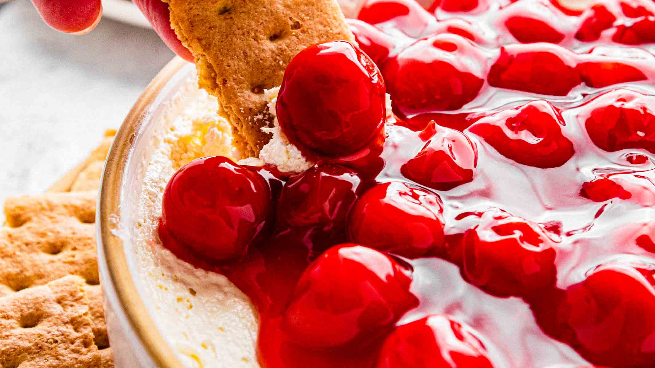 cherry cheesecake dip with cherry being lifted out of the bowl by the cracker