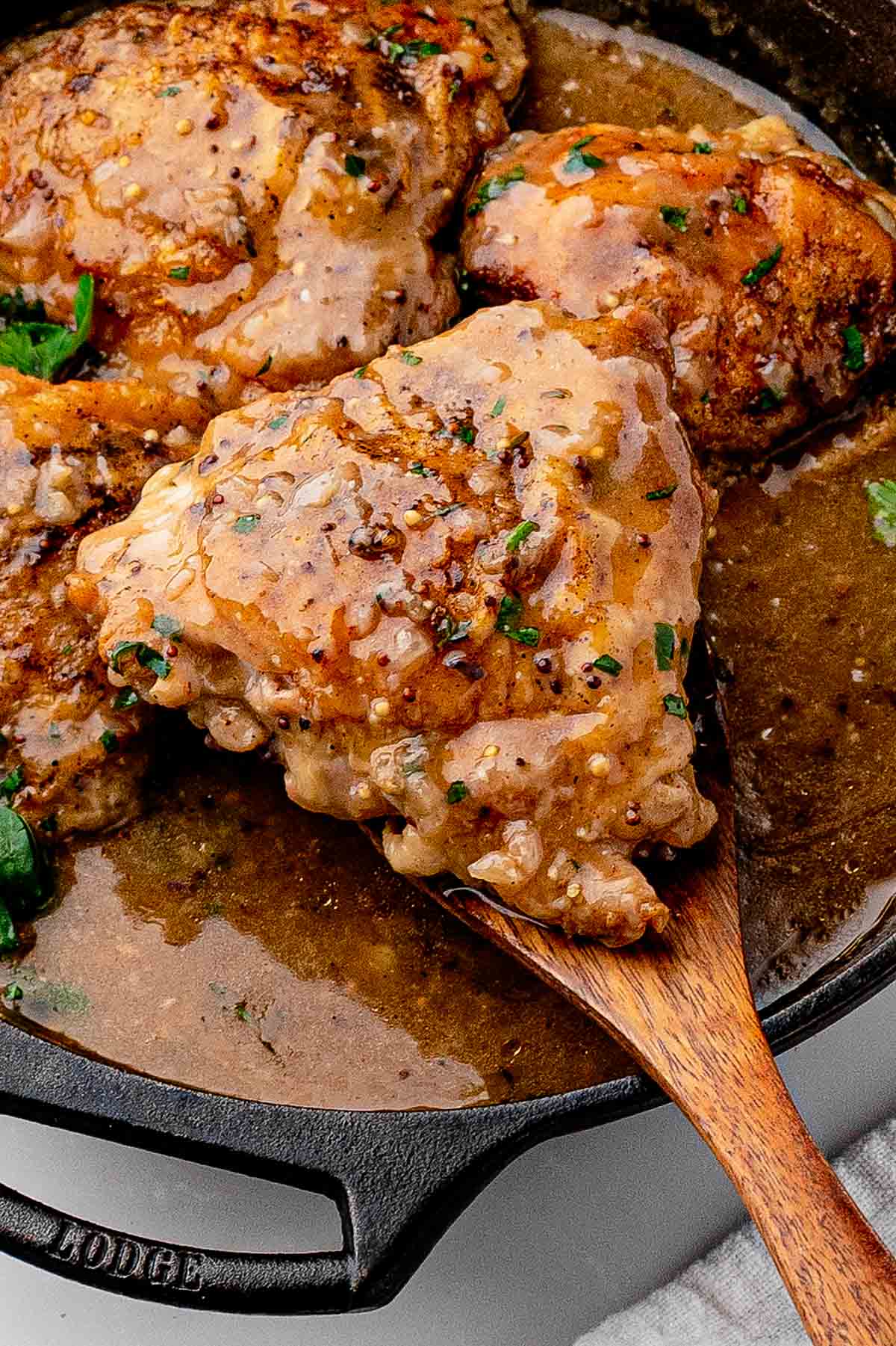 Wooden spoon coated in glossy beer braising sauce, held over a skillet of braised chicken thighs