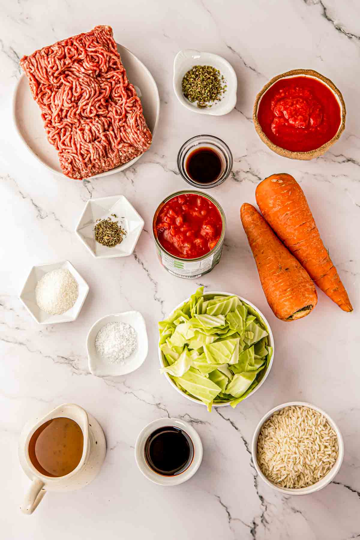 Ingredients for cabbage roll soup laid out on a counter: ground pork, cabbage, carrots, tomatoes, marinara, broth, and rice